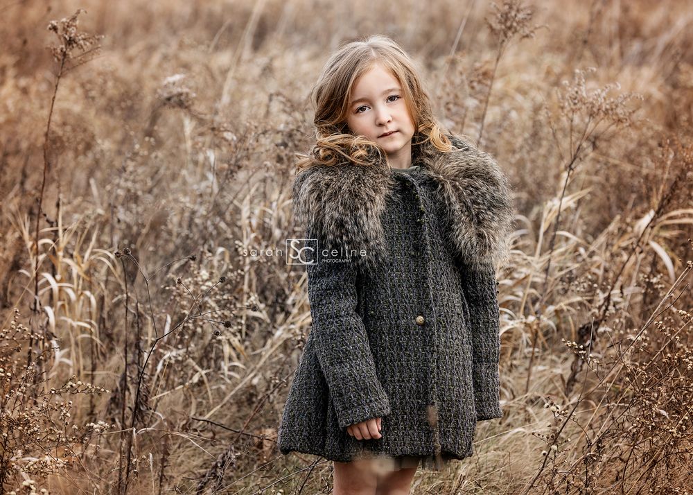 A child in a grey winter coat stands among tall dried grass in an autumn field.
