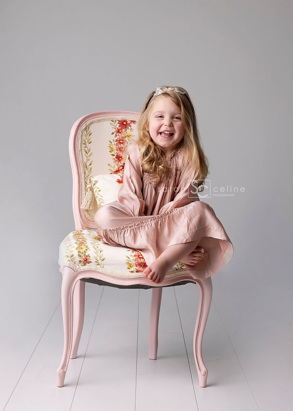 Joyful child in pink dress sitting on vintage floral chair against grey backdrop.