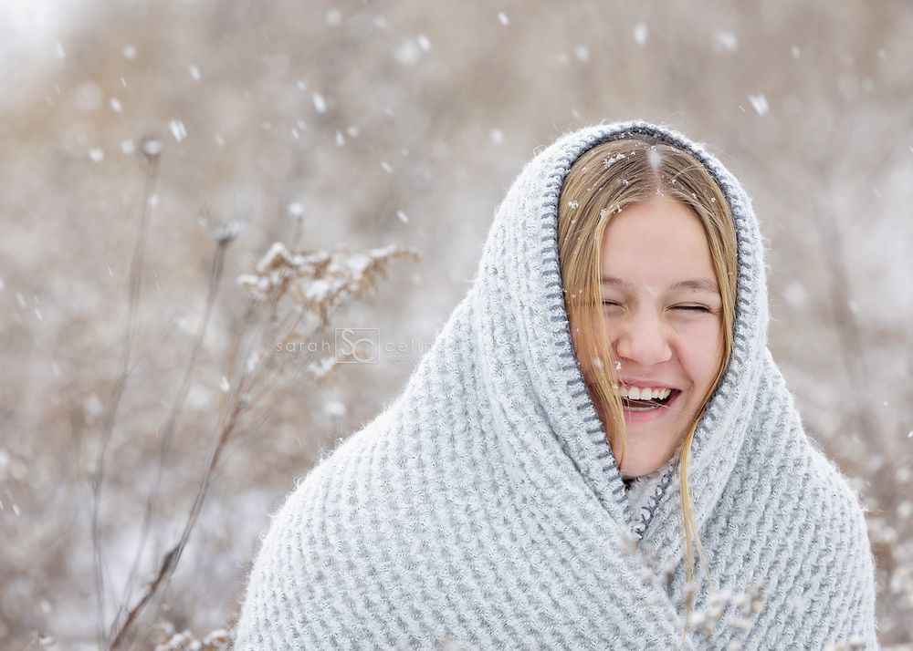 Candid winter portrait of someone wrapped in white knit blanket laughing in snowy setting.