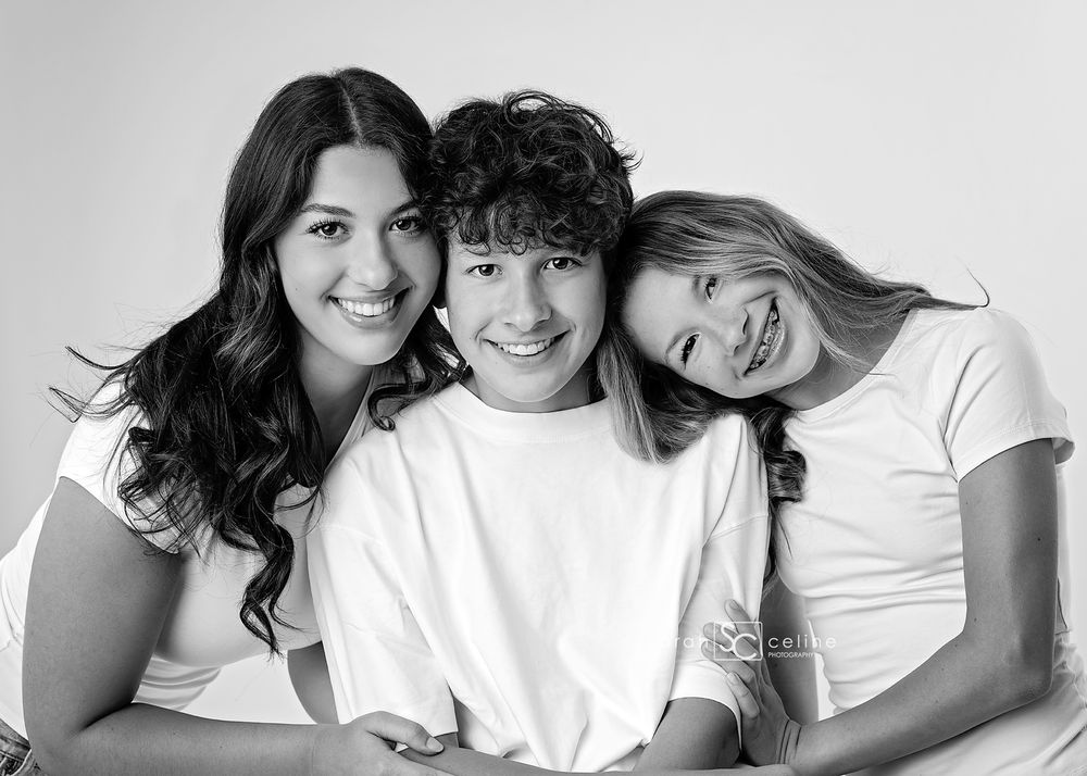 Black and white portrait of three smiling siblings in white shirts posing together for a professional family photo.