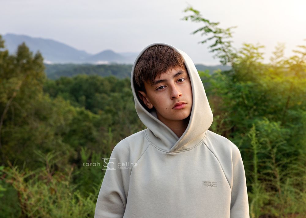 Person in cream hoodie poses in natural outdoor setting with mountains and greenery in background at golden hour.