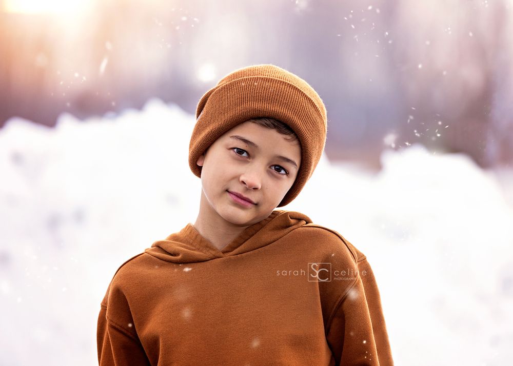 A person wearing a brown beanie and matching hoodie stands in a snowy winter scene with soft lighting.