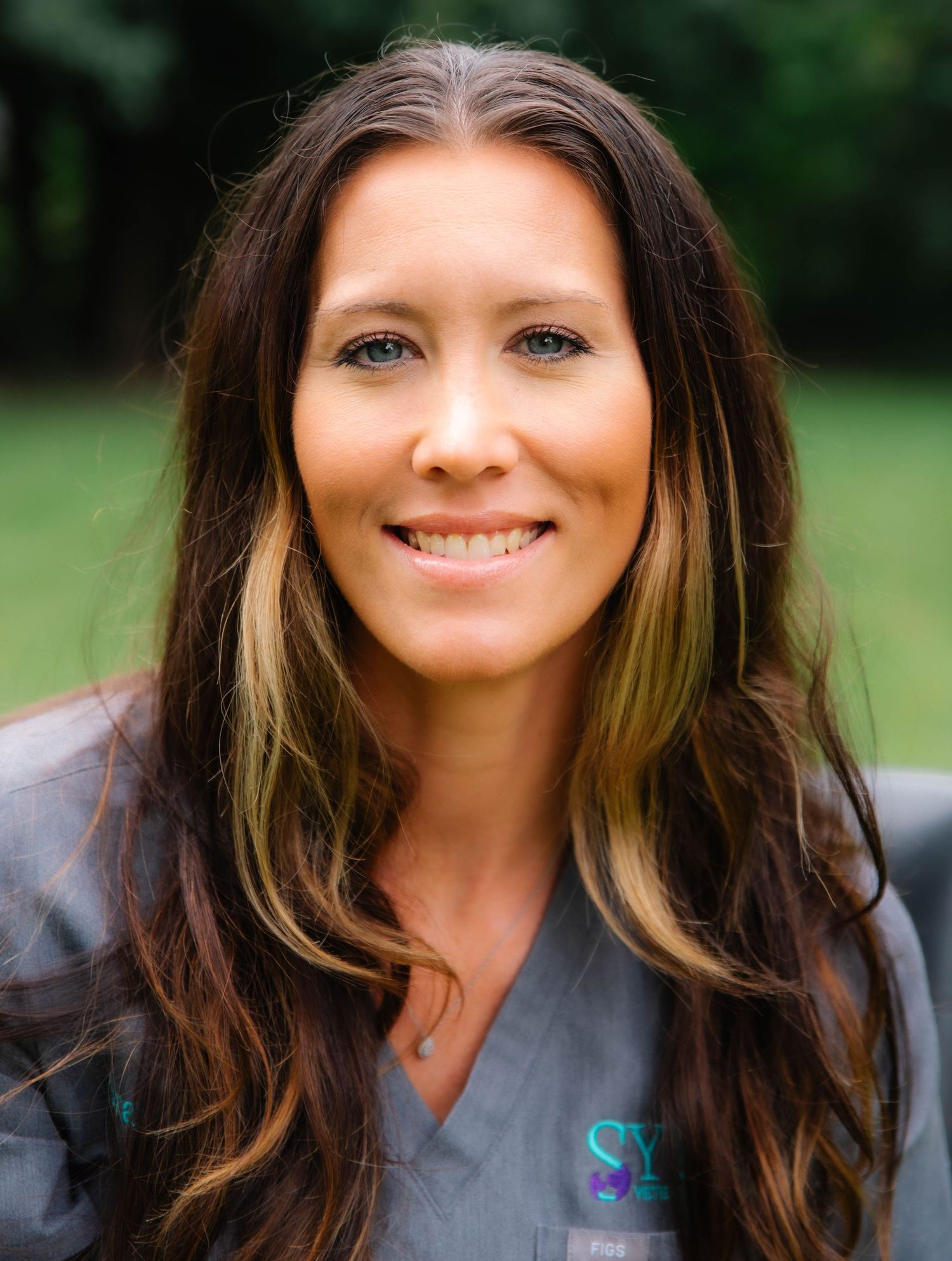 Smiling female healthcare professional with long brown highlighted hair wearing gray medical scrubs outdoors.