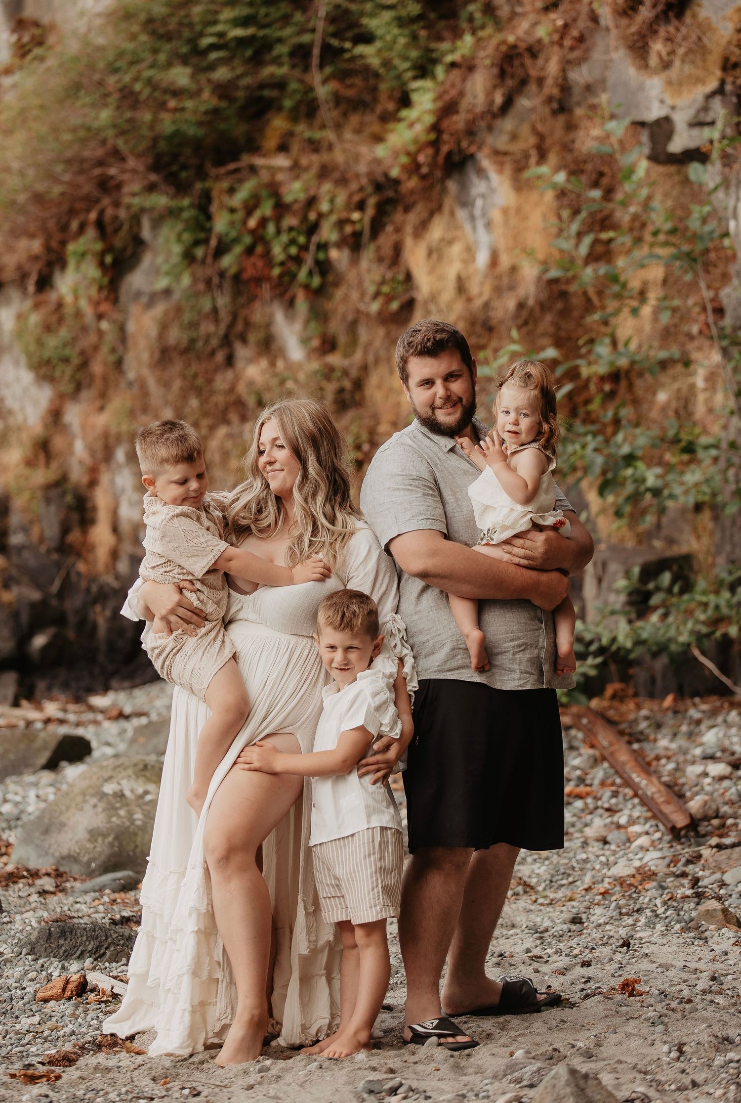 Pregnant woman in flowing white dress poses with husband and three young children on a rocky beach with autumn foliage.