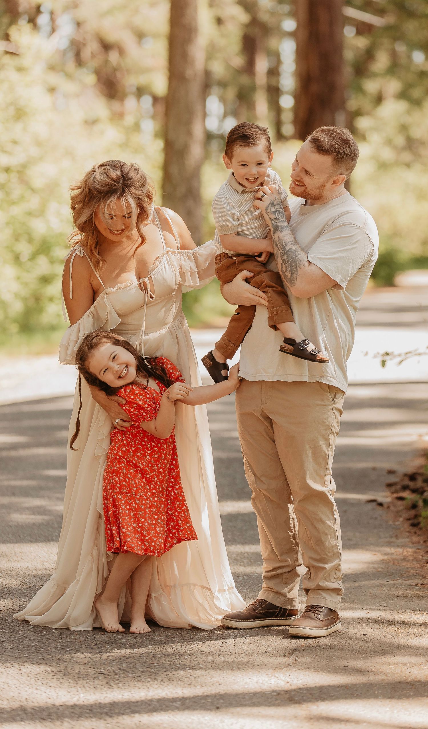 Happy family of four posing outdoors, mom in white dress, dad holding toddler, young girl in red dress smiling.