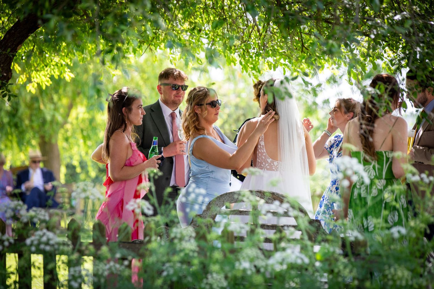 Guests mingle outdoors at a summer wedding ceremony surrounded by lush green trees and wildflowers.