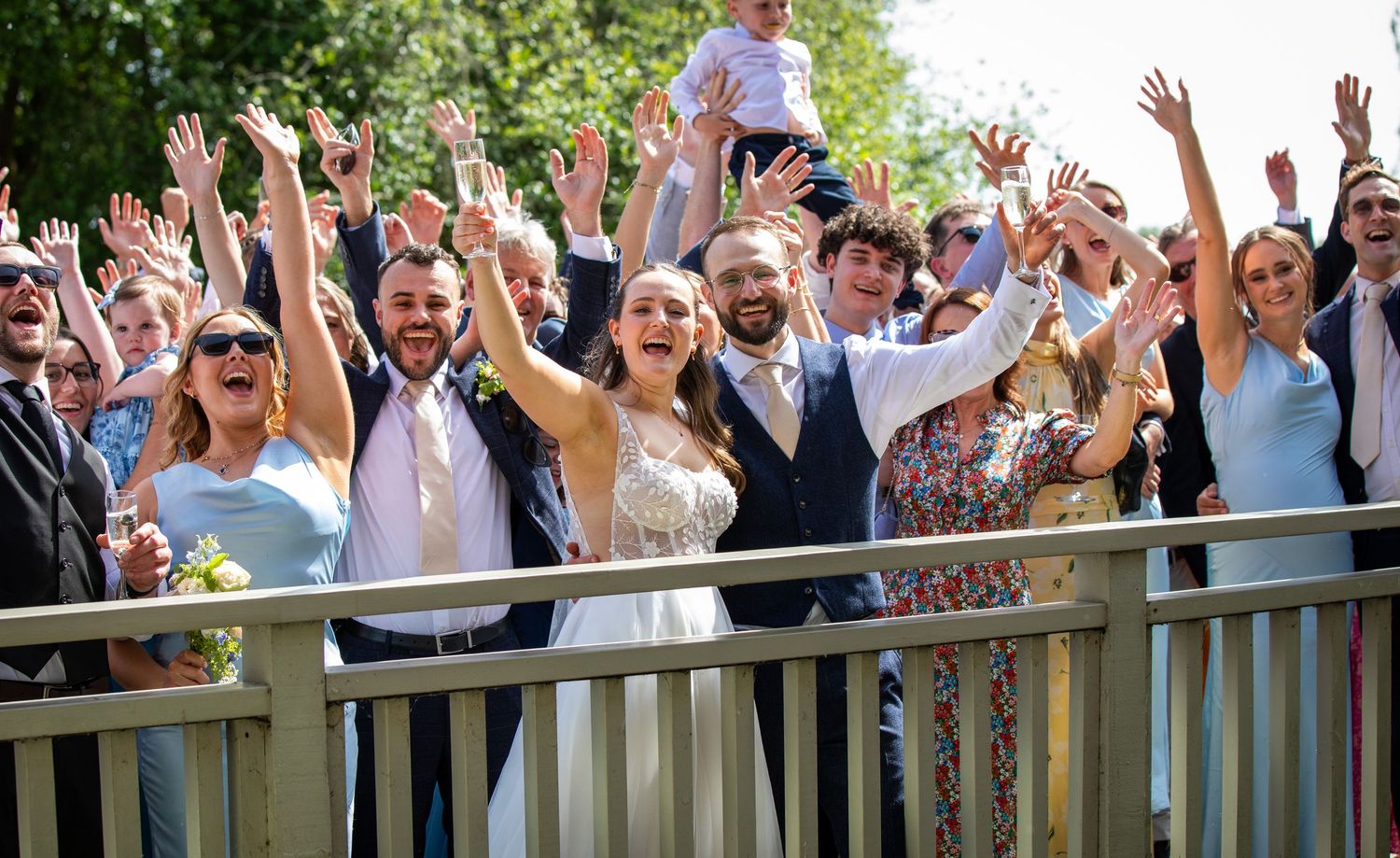 Wedding party celebrating on a bridge with raised arms, bride and groom centered among joyful guests on a sunny day.