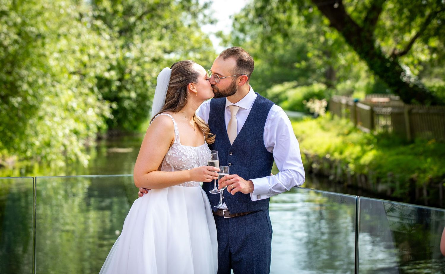 Bride and groom kissing while holding champagne glasses outdoors near a river with lush green trees.