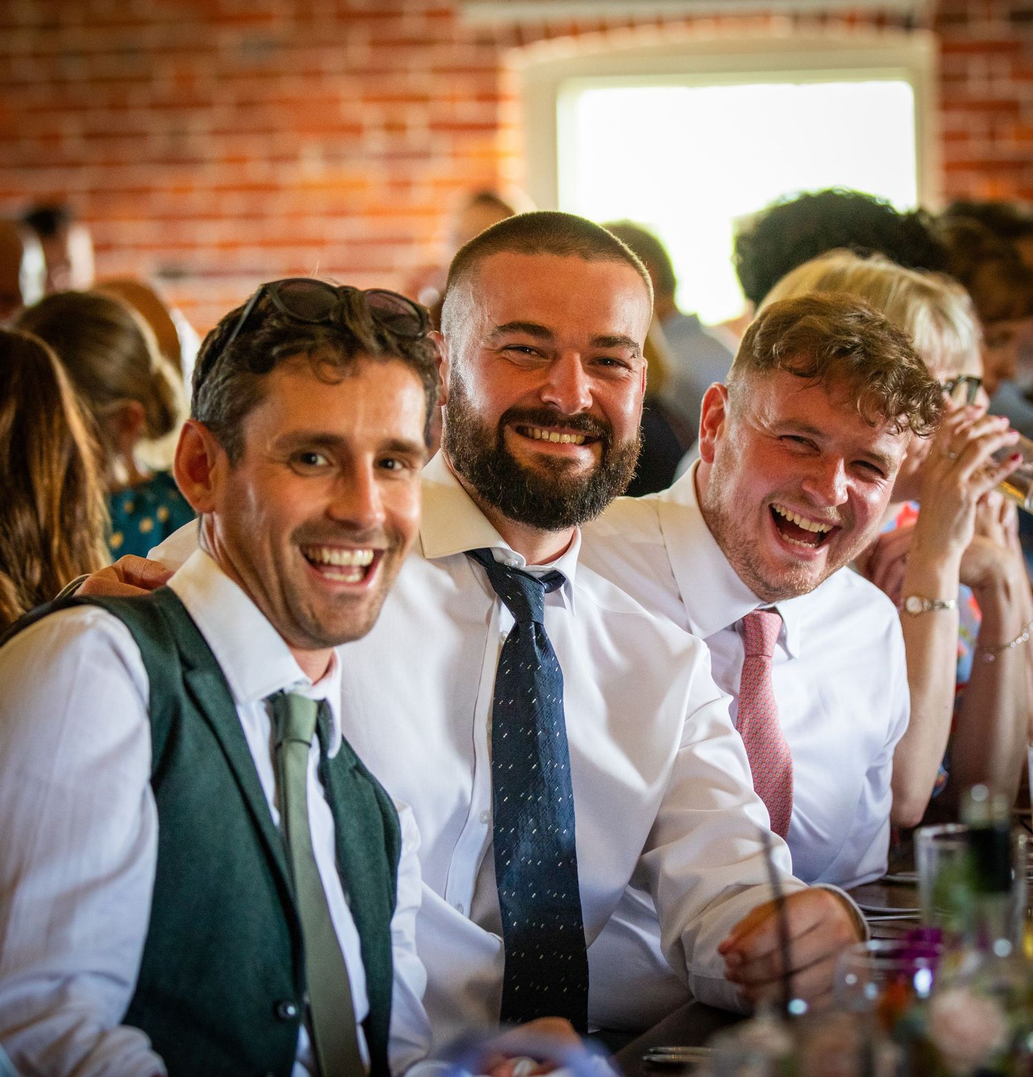 Three smiling men in dress shirts and ties pose together at a wedding reception dinner.