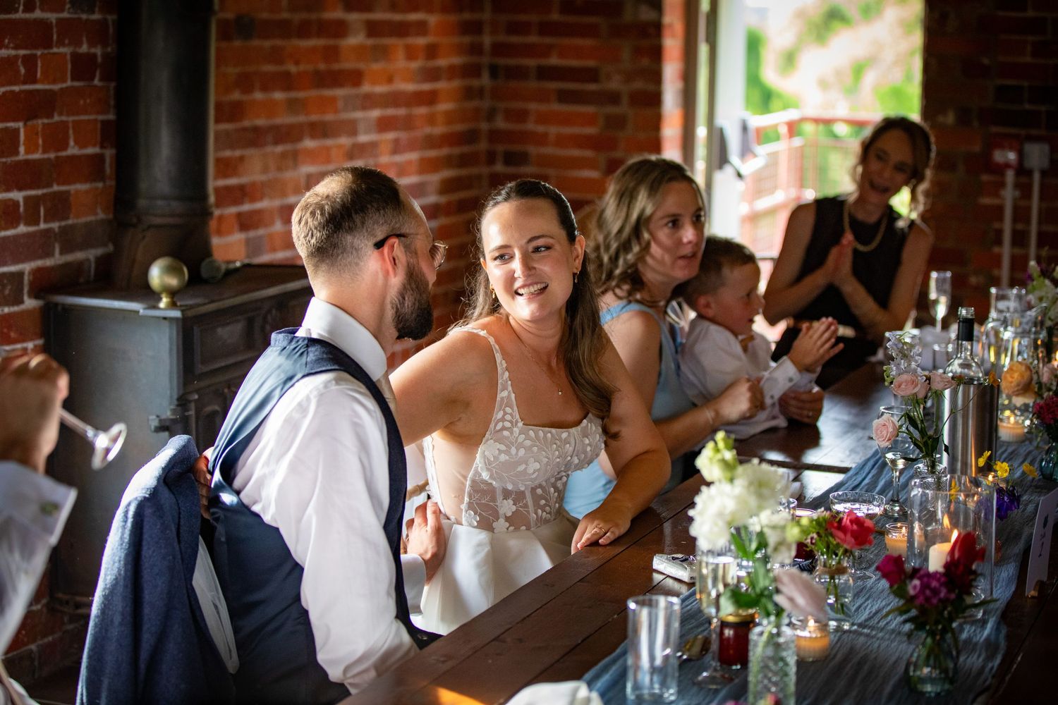 Bride smiling at groom during wedding reception at rustic brick venue with colorful floral table decorations.