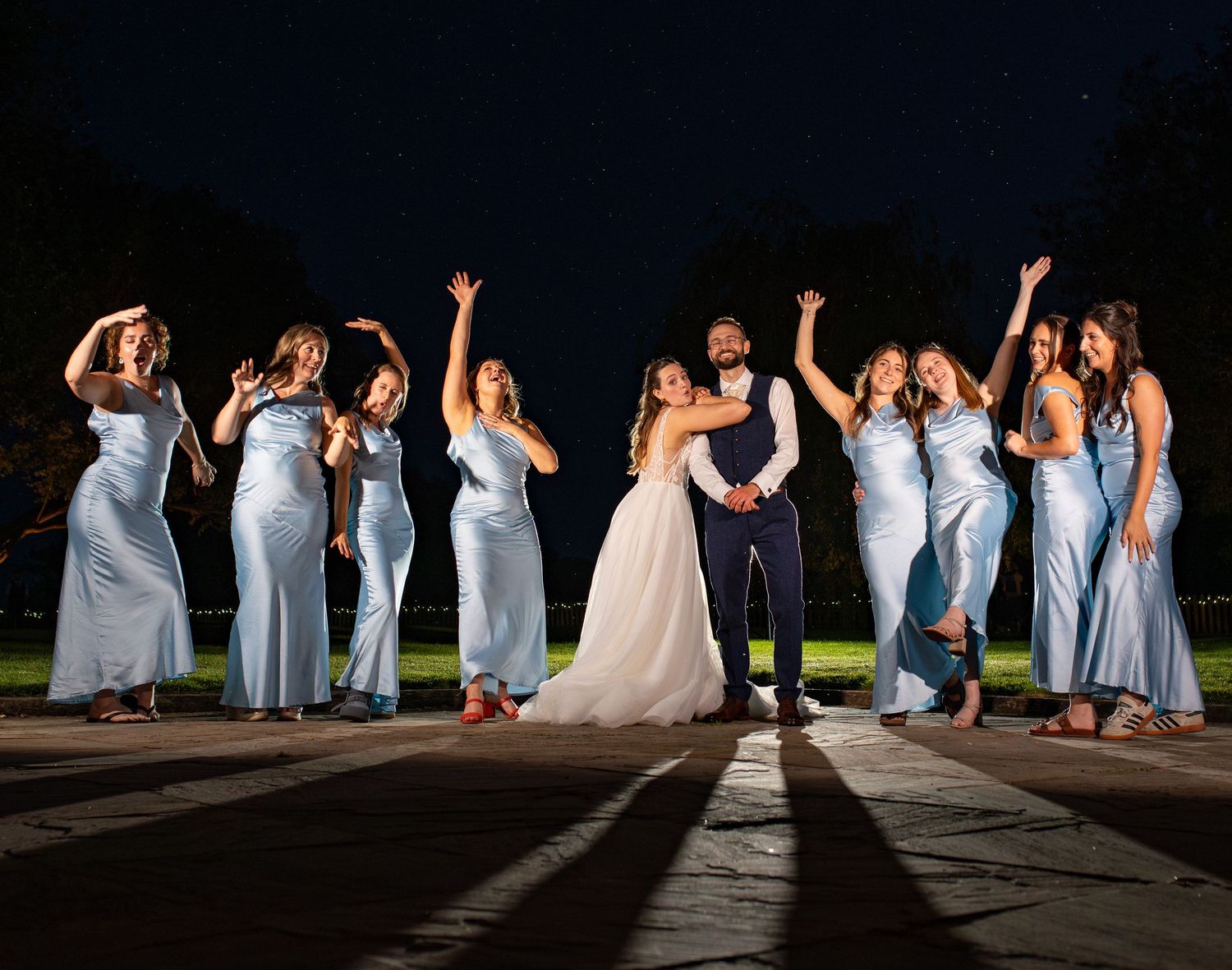Bride and groom celebrate with bridesmaids in blue satin gowns, arms raised joyfully at an outdoor night wedding.