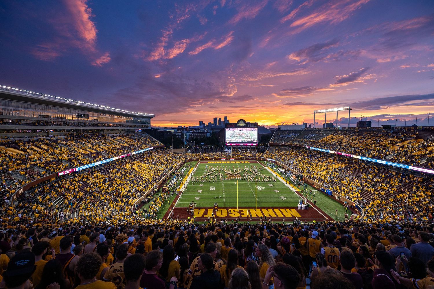 Camera looking into a full stadium at sunset and marching band marching a block M down the field.