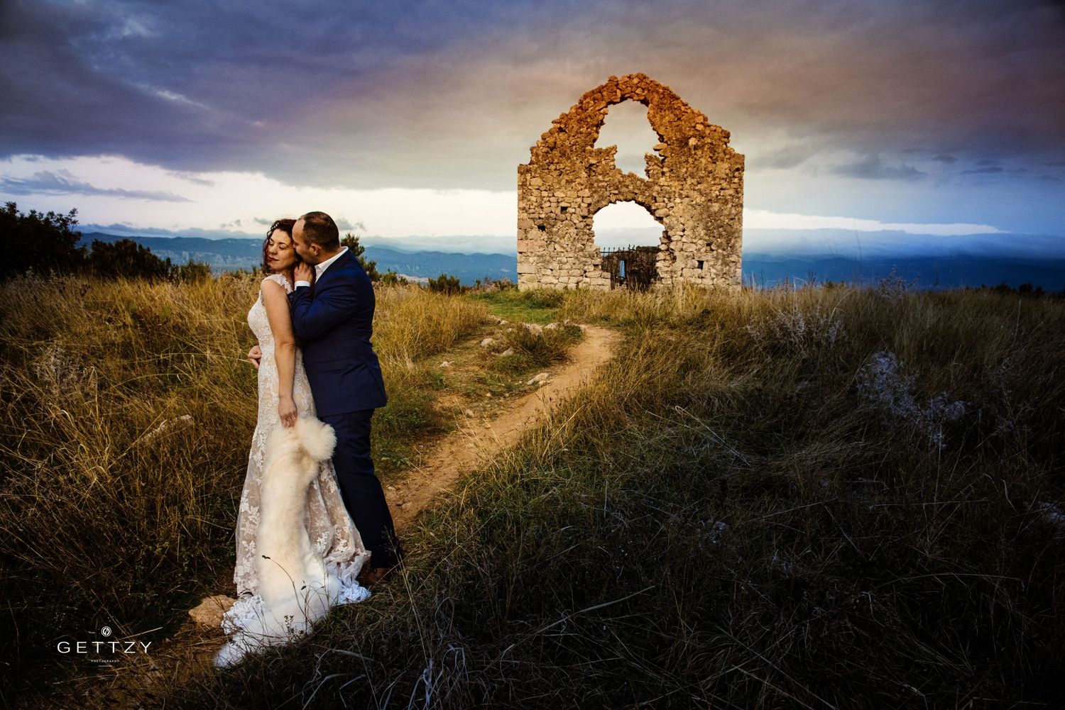 A couple in wedding attire embrace near ancient stone ruins at sunset on a hilltop overlooking a dramatic landscape.