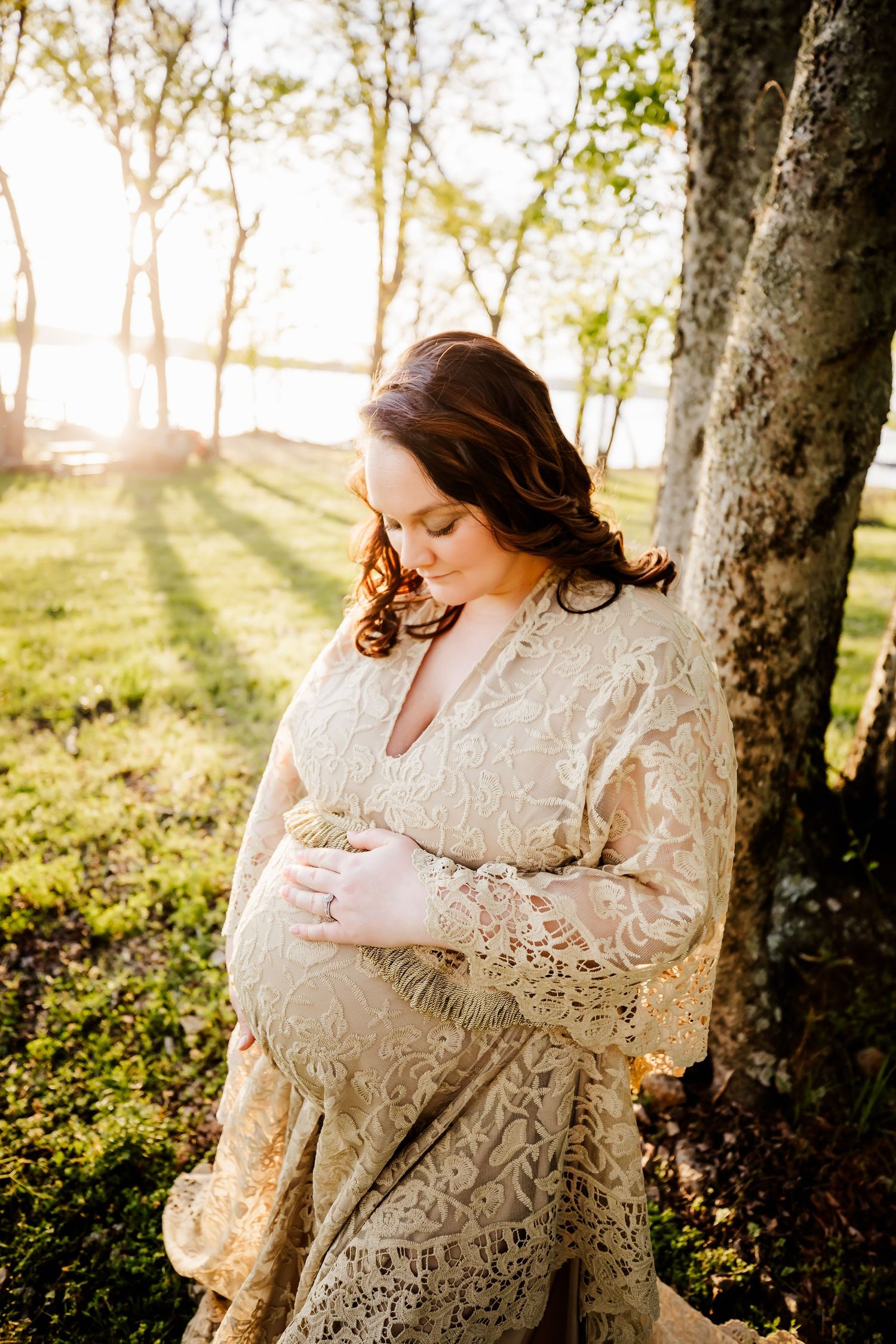Pregnant woman in white gown smiling and holding belly at sunset next to tree in Dickson, TN
