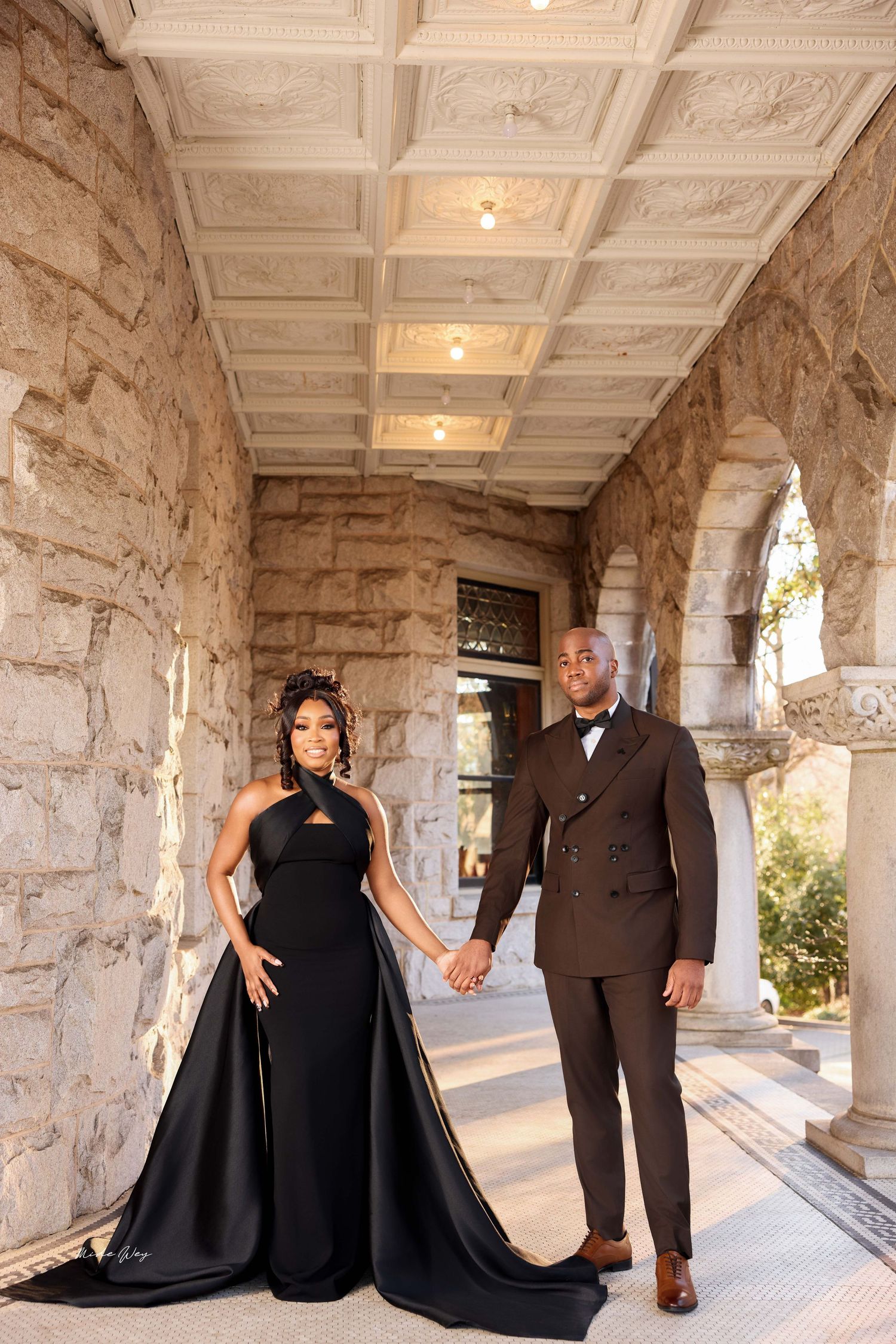 A romantic couple poses for photos in a stone archway corridor with elegant black and brown formal attire.