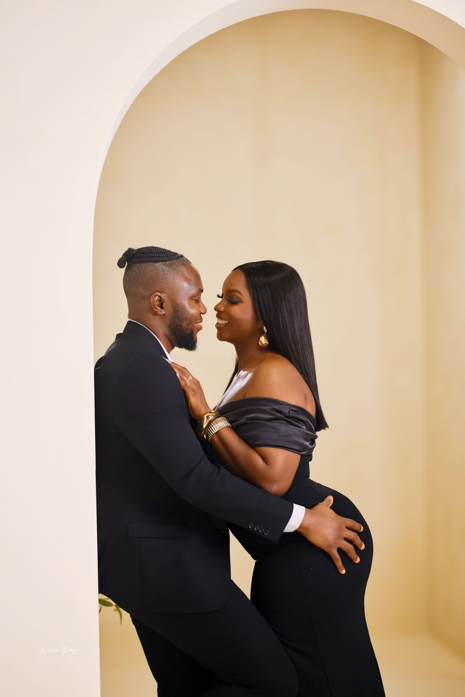 Romantic couple in matching black outfits embrace intimately against beige wall with arched alcove.
