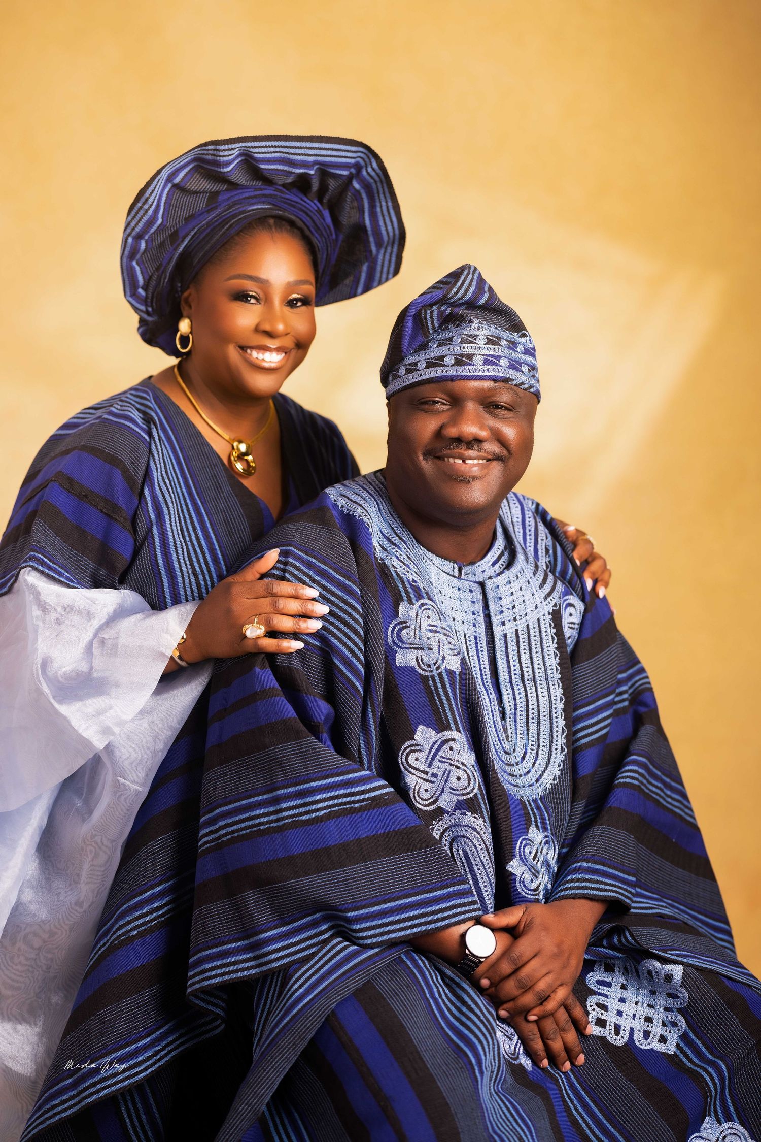 A couple wearing traditional blue and white African attire poses together in a warm-toned studio portrait series.