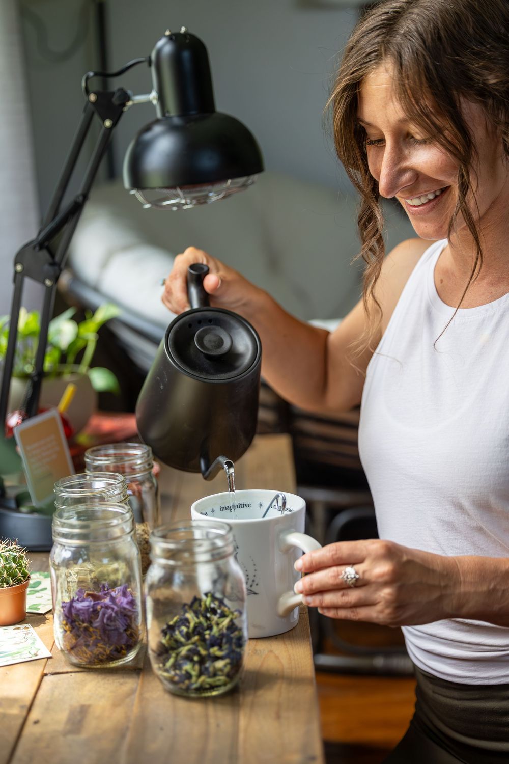 Person in white tank top preparing herbs and ingredients in glass jars under desk lamp.