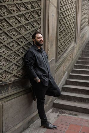 Person in black attire posing against ornate architectural wall with geometric patterns.
