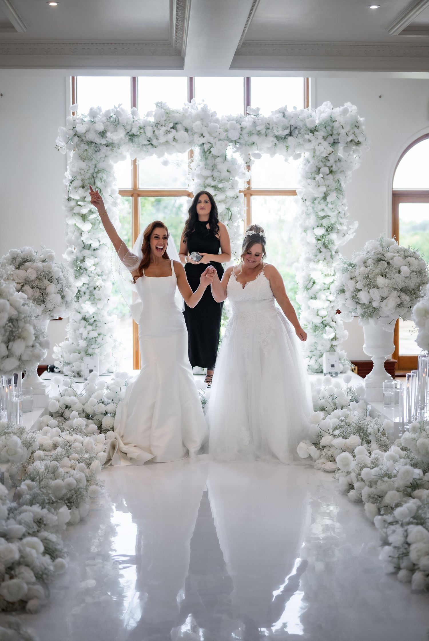 Two brides celebrate with a celebrant under an elaborate white floral arch in a sunlit ceremony space.