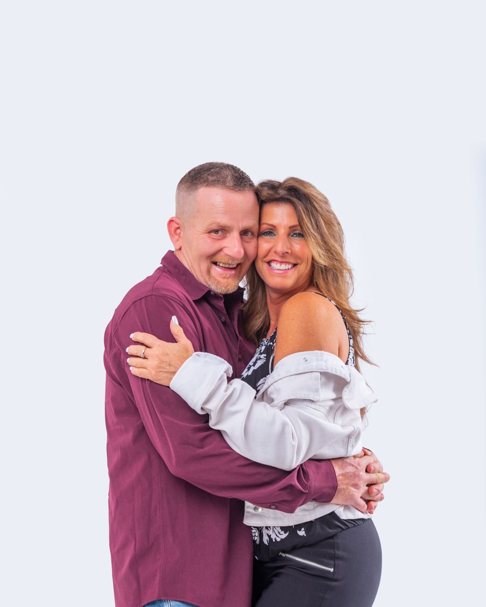 A loving couple embraces while smiling against a bright background, wearing burgundy and white casual attire.