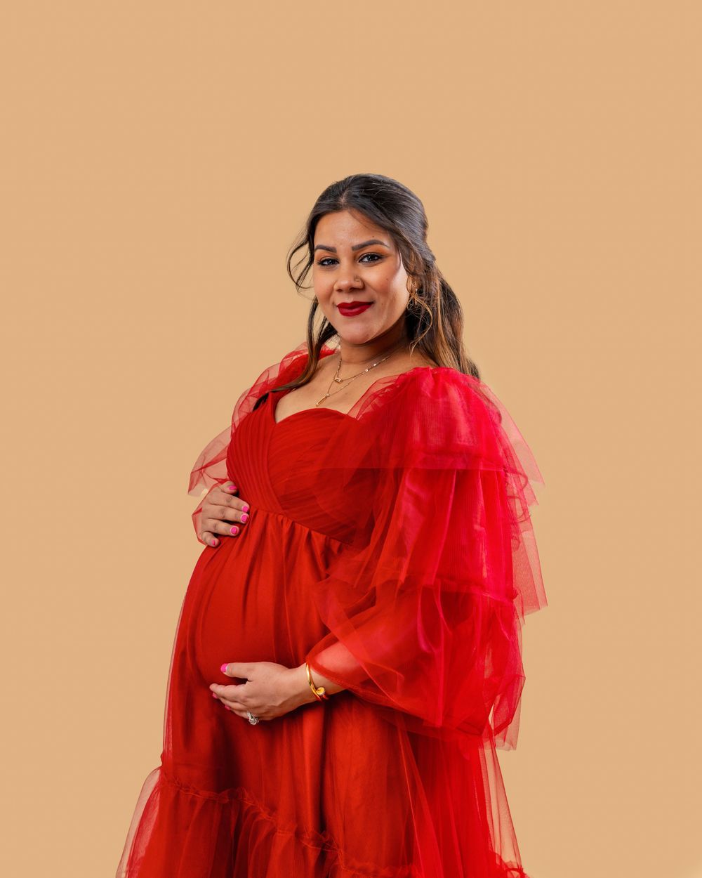A woman in a vibrant red tulle maternity gown poses against a beige backdrop with hands on her baby bump.