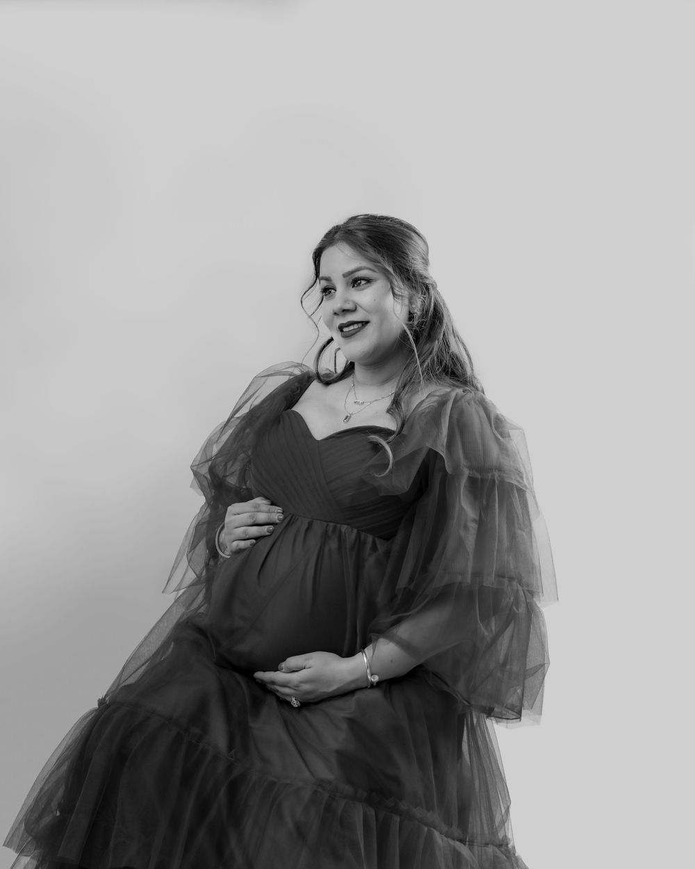A smiling expectant mother in a flowing black tulle dress poses for a maternity photo against a plain backdrop.