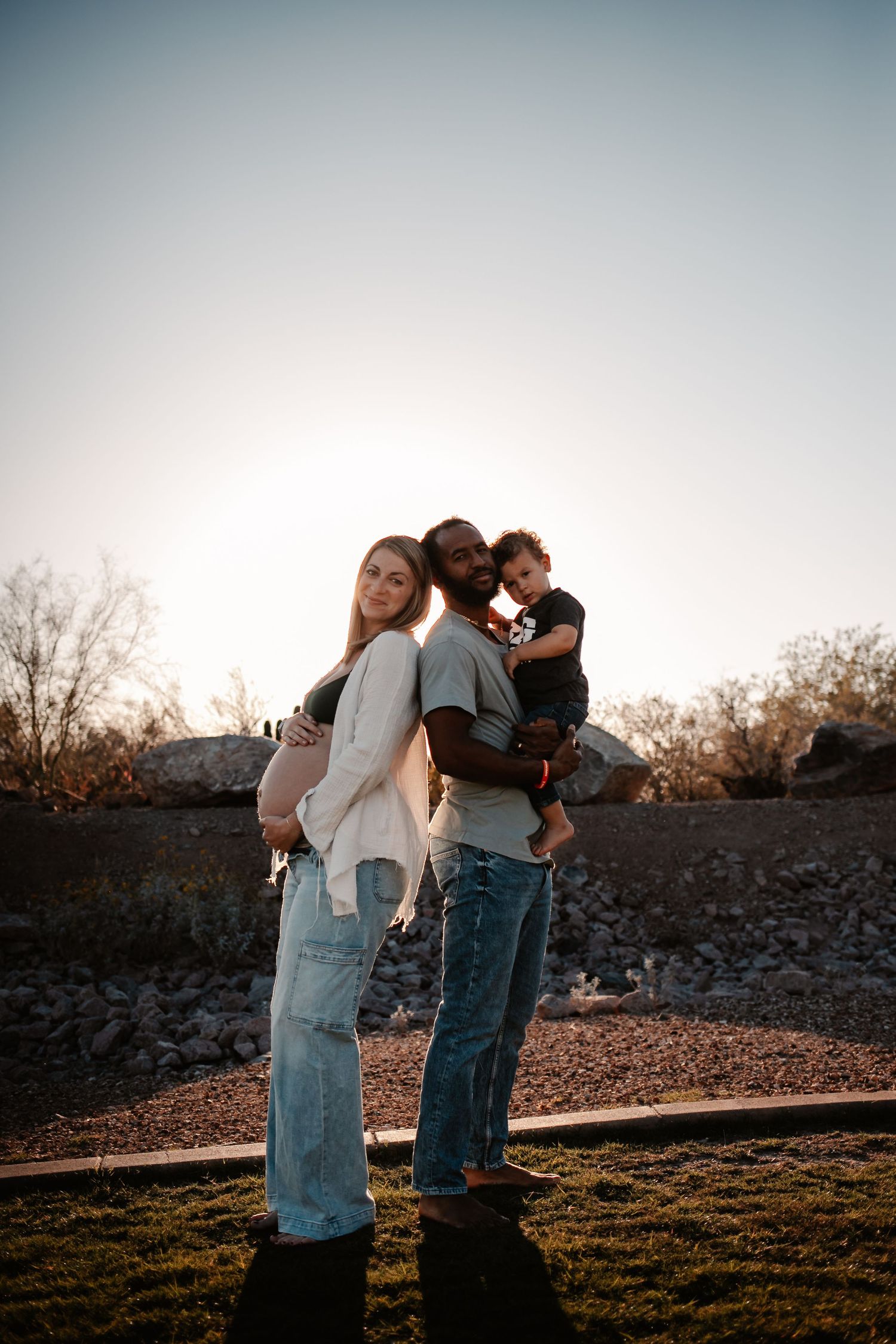 Pregnant woman and man holding toddler stand back-to-back outdoors at golden hour in jeans.