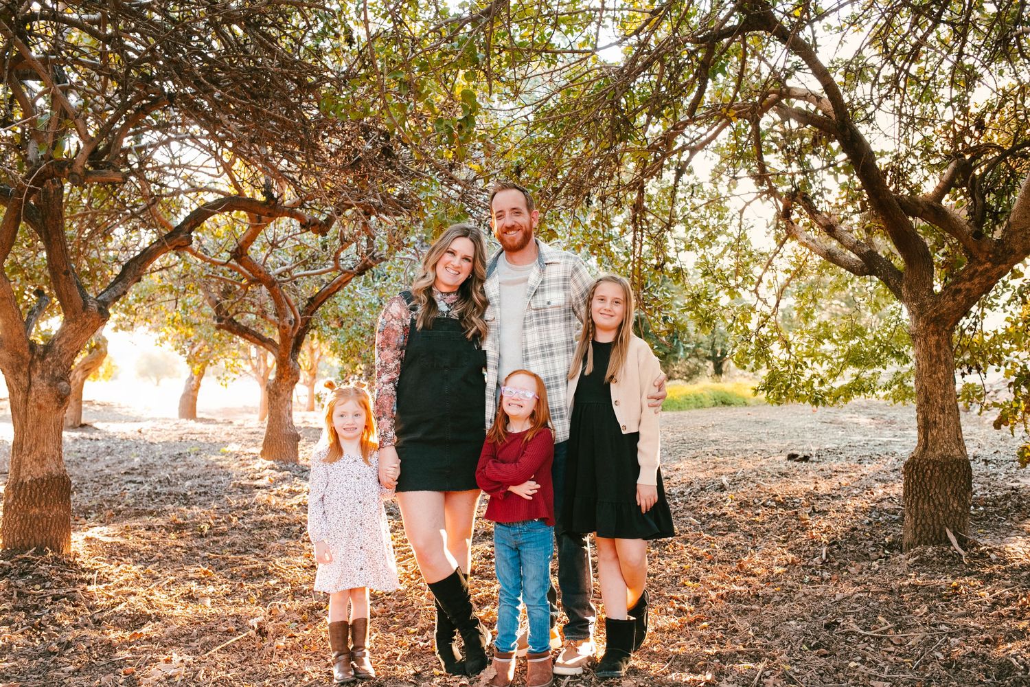 Family of five posing outdoors among autumn trees in warm golden light, with two young girls and a teenager smiling.