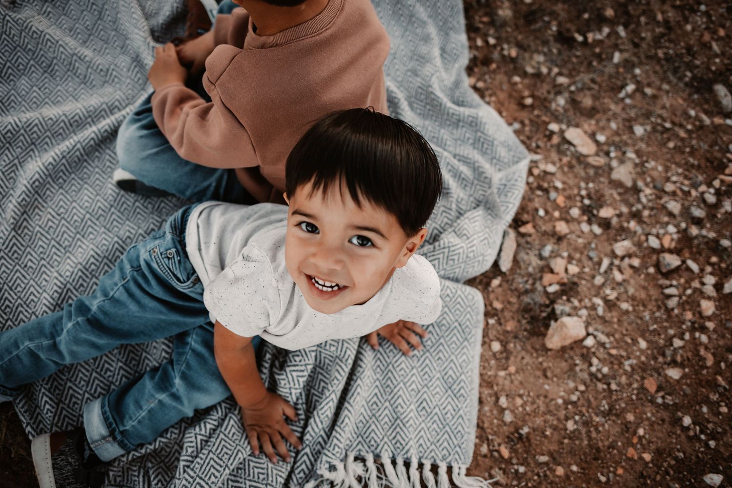Smiling toddler boy with dark hair wearing white shirt and jeans, wrapped in gray blanket outdoors.