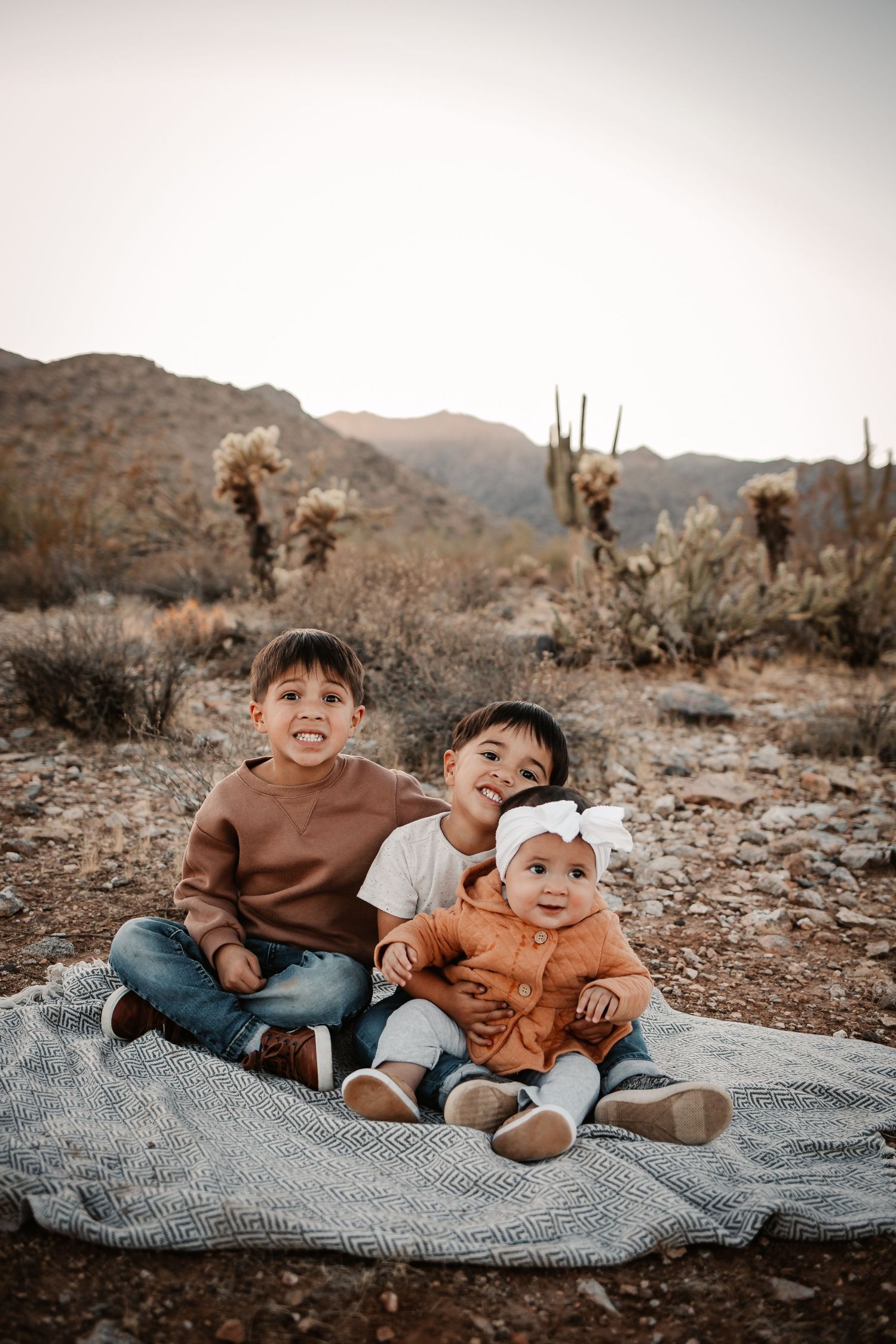 Three young siblings sit on a blanket in a desert landscape with cacti and mountains in the background.