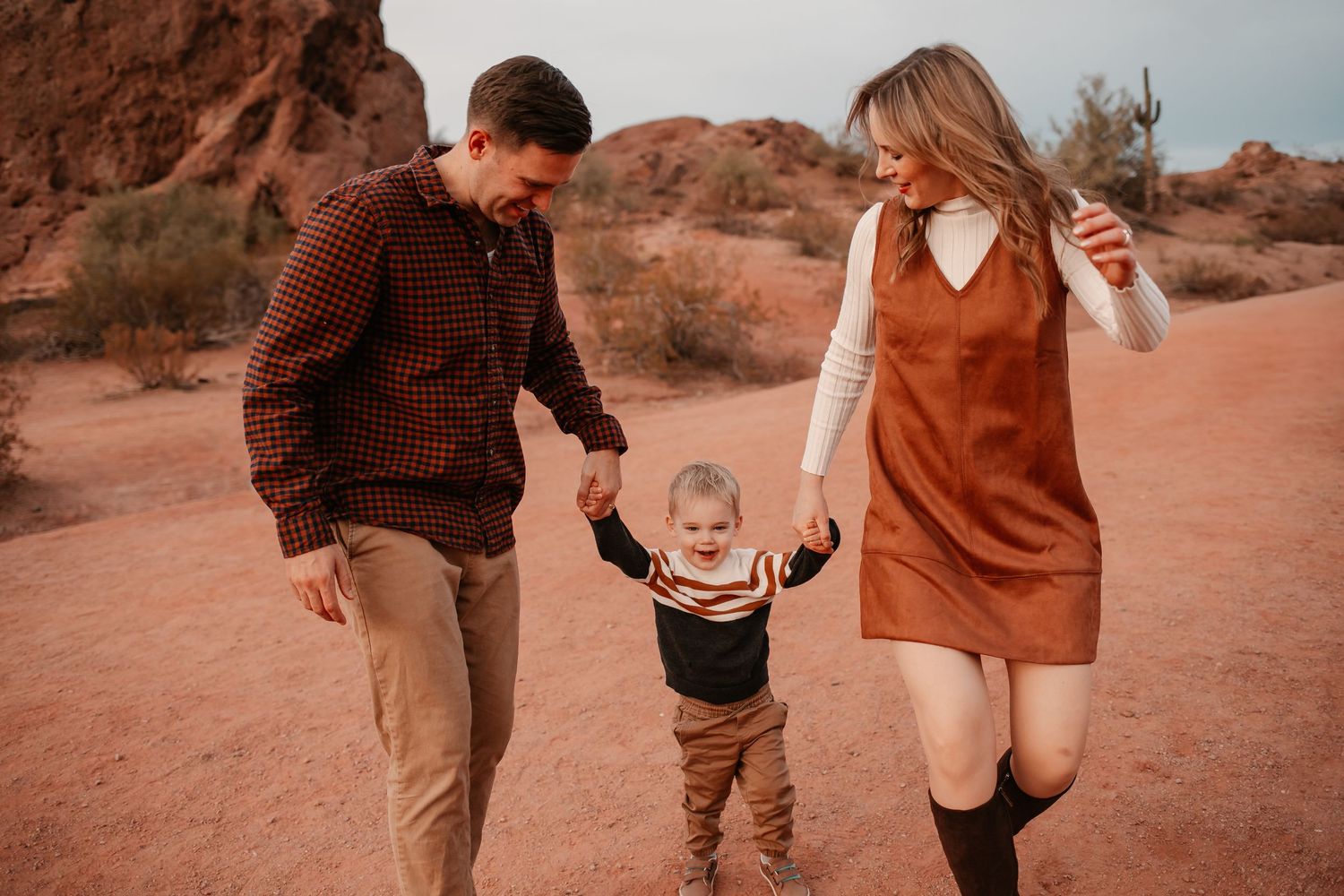 A young family walks hand-in-hand with their toddler in a scenic desert landscape with red rock formations.