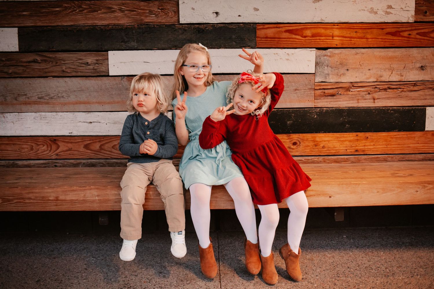 Three young children sitting on a bench smiling and posing in front of a rustic wooden plank wall.