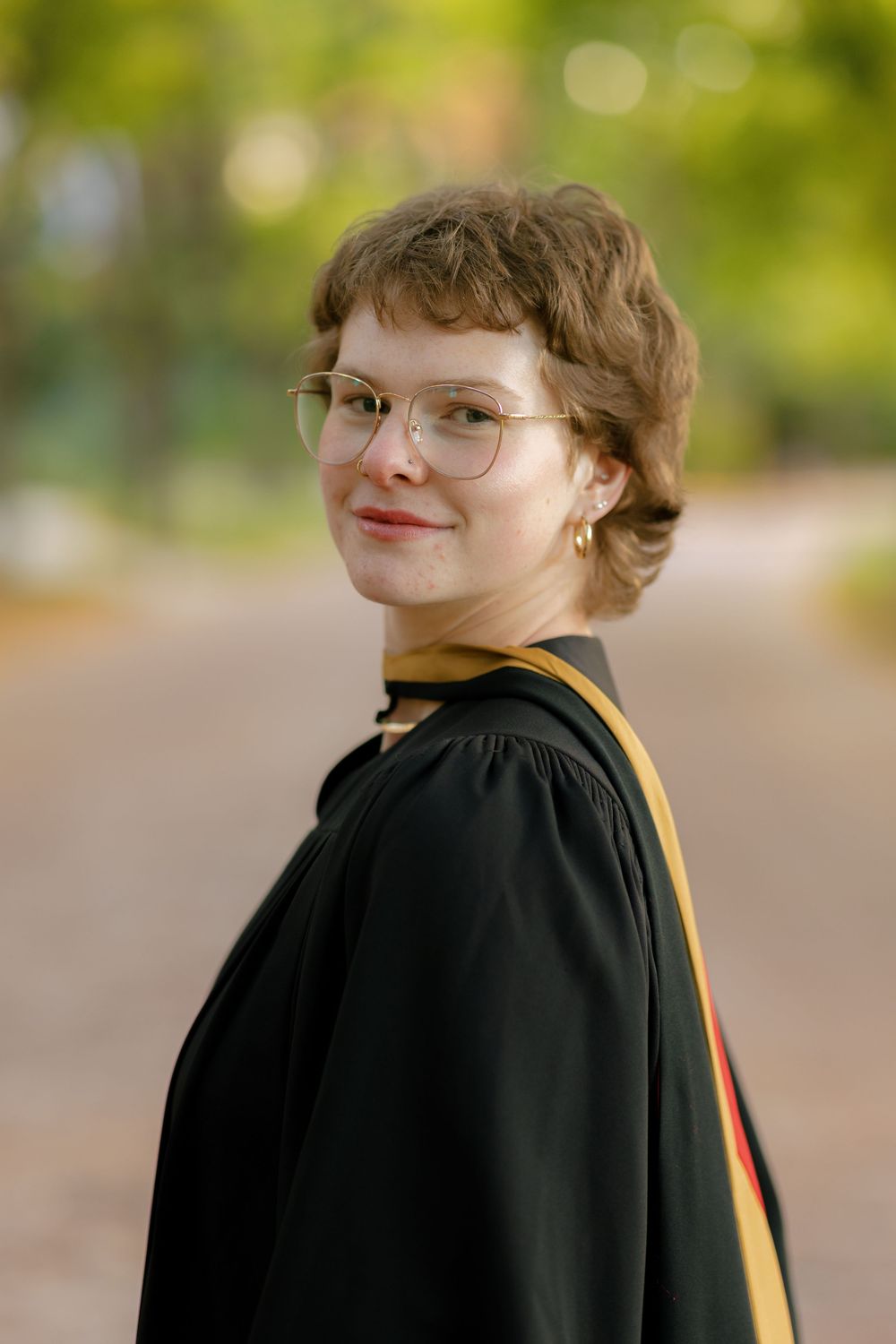 A person wearing a black graduation gown and glasses smiles while looking over their shoulder outdoors.