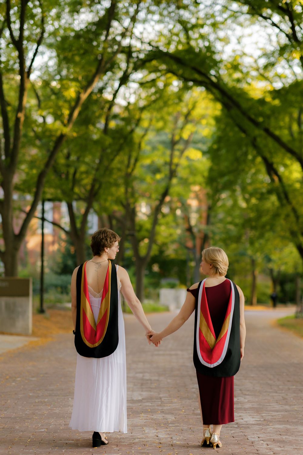 Two people holding hands while walking down a tree-lined path wearing colorful draped tops and contrasting skirts.