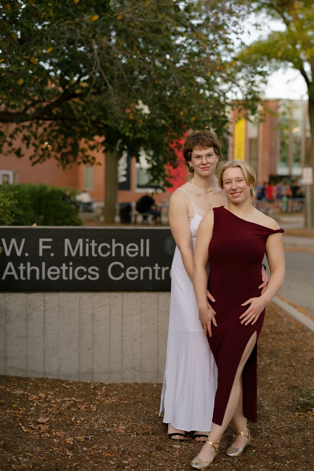 Two dancers pose together in formal attire outside the W.F. Mitchell Athletics Centre during autumn.