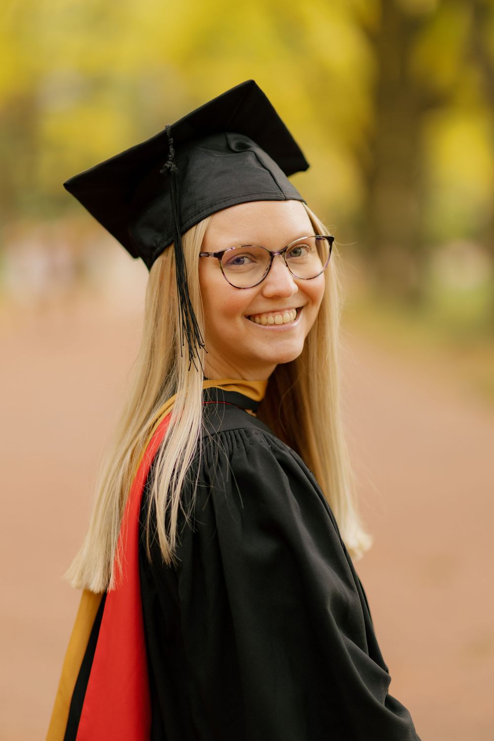 A graduate in cap and gown smiles while posing outdoors with autumn foliage in the background.