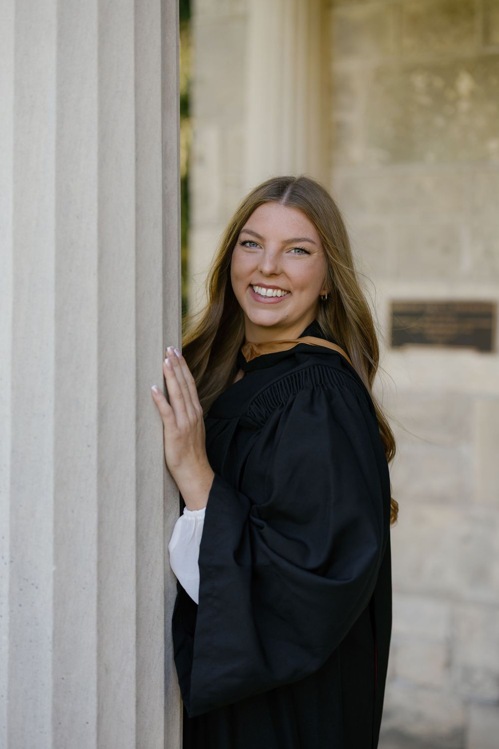 A graduate in a black academic gown smiles while leaning against a stone column outdoors.