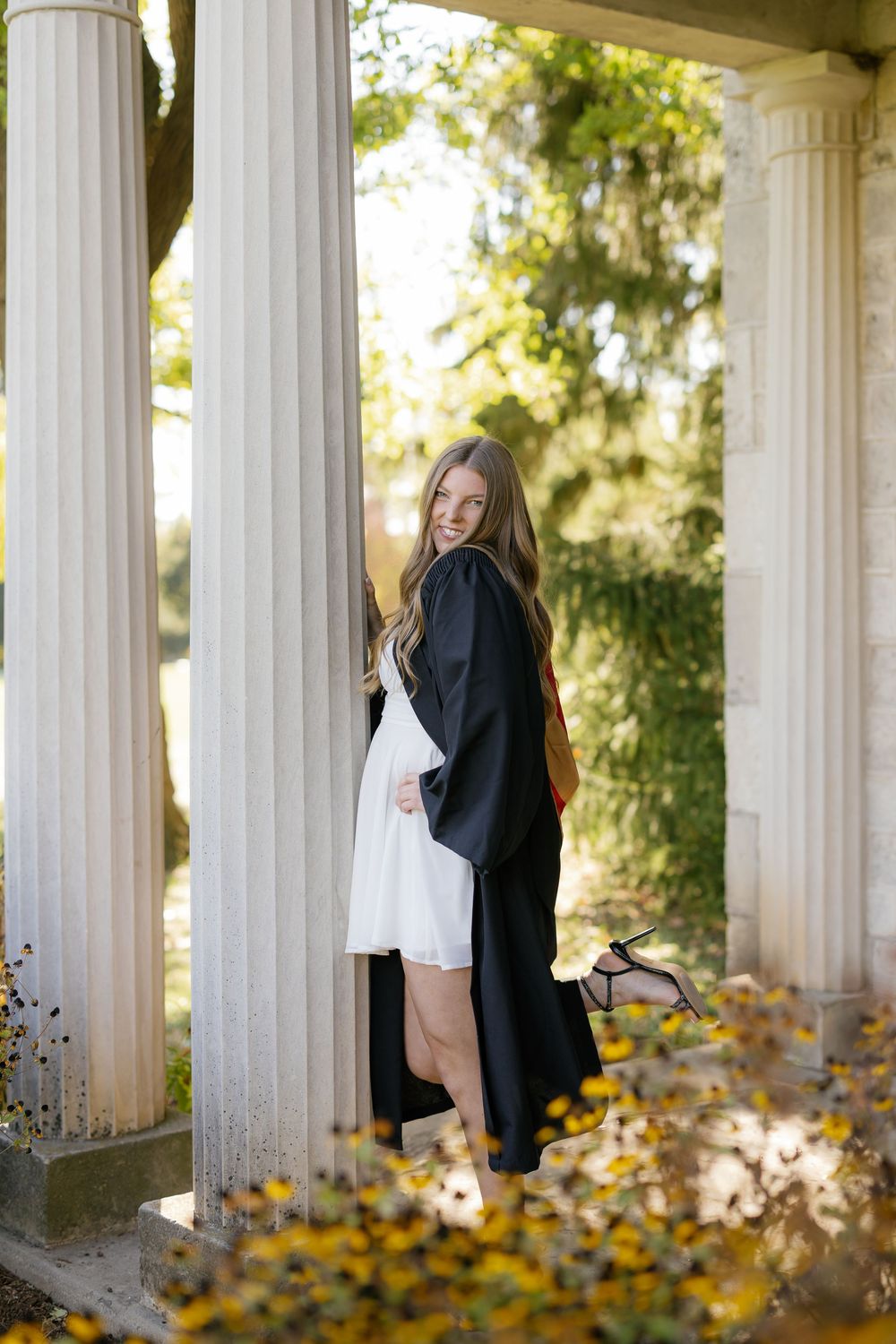 Graduate in black cap and gown poses between white columns with autumn leaves scattered on ground.