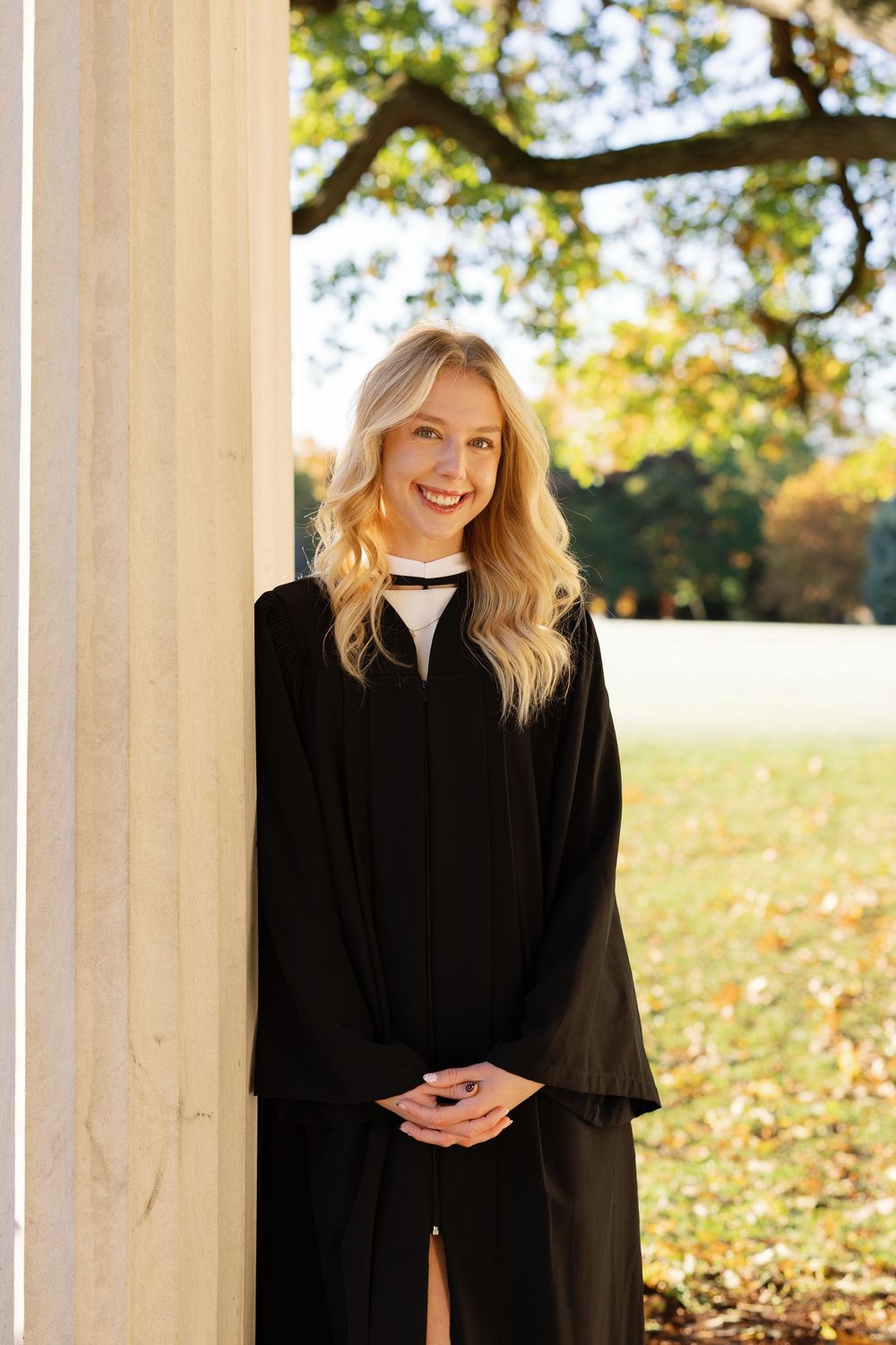 A person in a black graduation gown stands in front of white columns on an autumn day with fallen leaves.