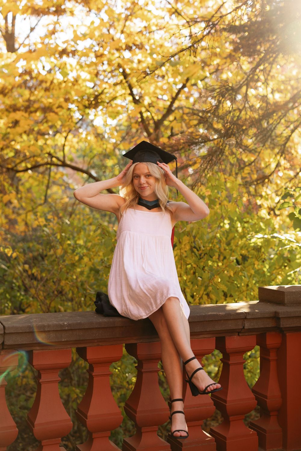 A person in a white dress poses playfully on a wooden railing with autumn foliage in the background.