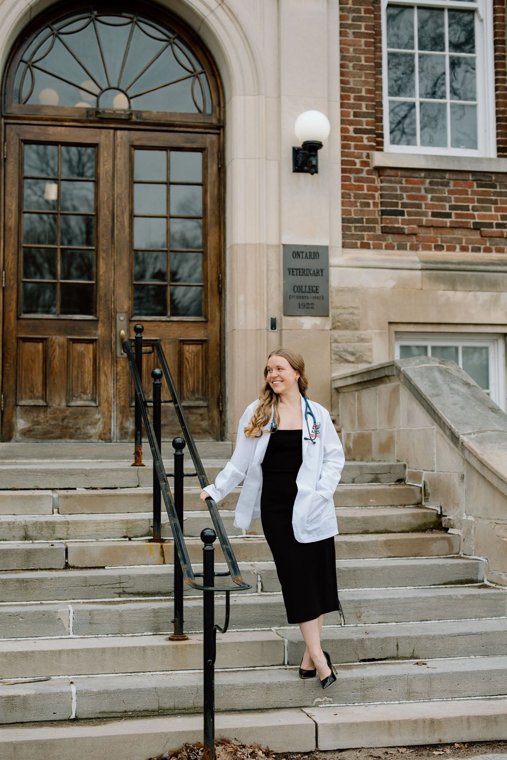 A woman in a white denim jacket and black dress poses on stone steps outside a brick building with arched doorways.