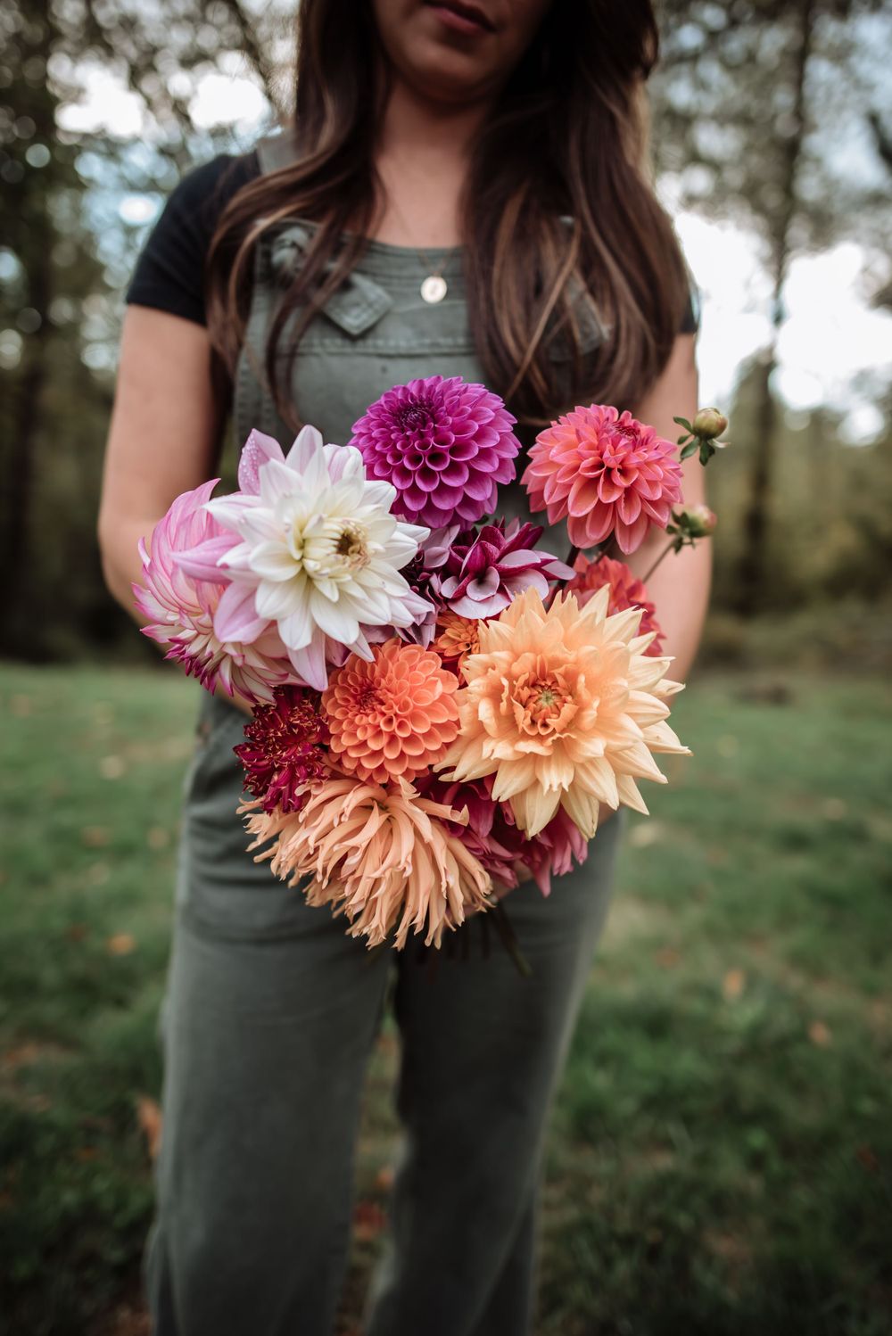 Person holding vibrant bouquet of pink, purple, and orange dahlias against forest backdrop.
