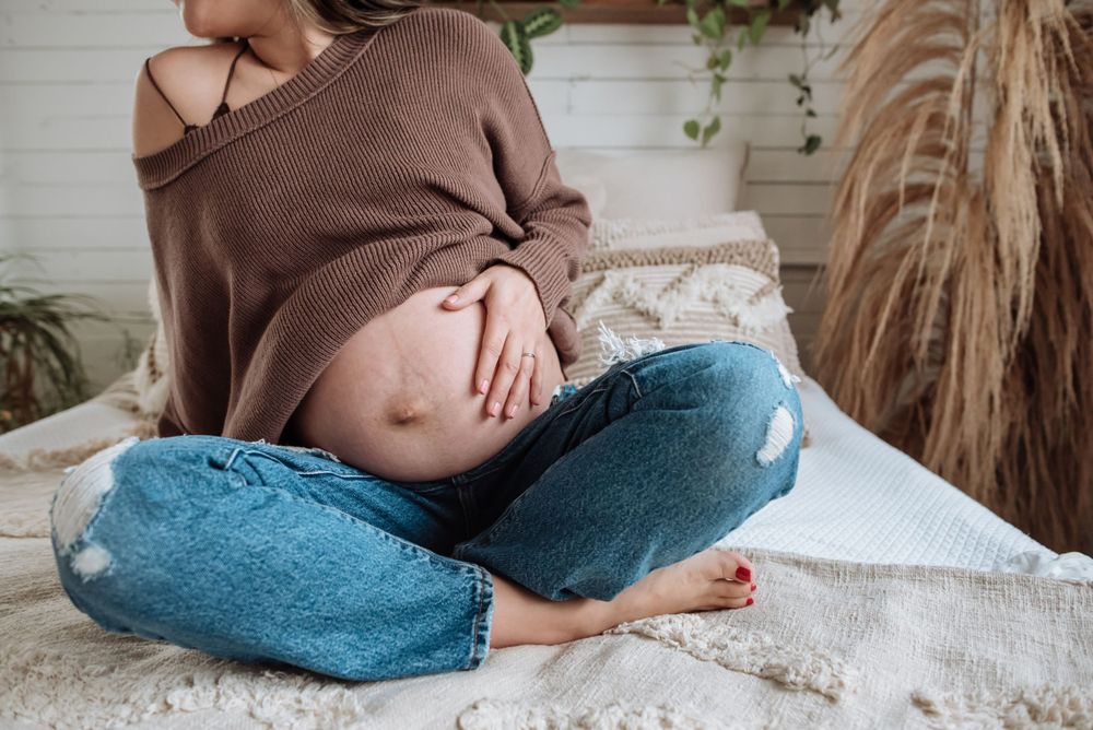 A pregnant woman in brown sweater and jeans sits cross-legged on white bedding.