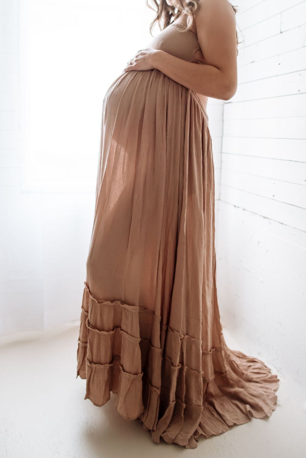 A side view of a pregnant woman wearing a flowing brown maxi dress in a bright white room.