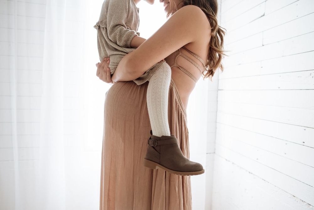 A tender moment captured showing a parent in a long beige dress holding a small child against white walls.