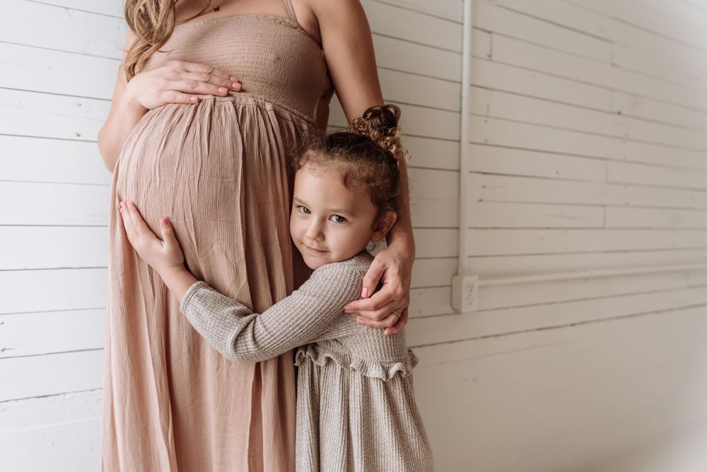 A touching maternity moment with a child embracing a pregnant belly while wearing a soft gray dress.