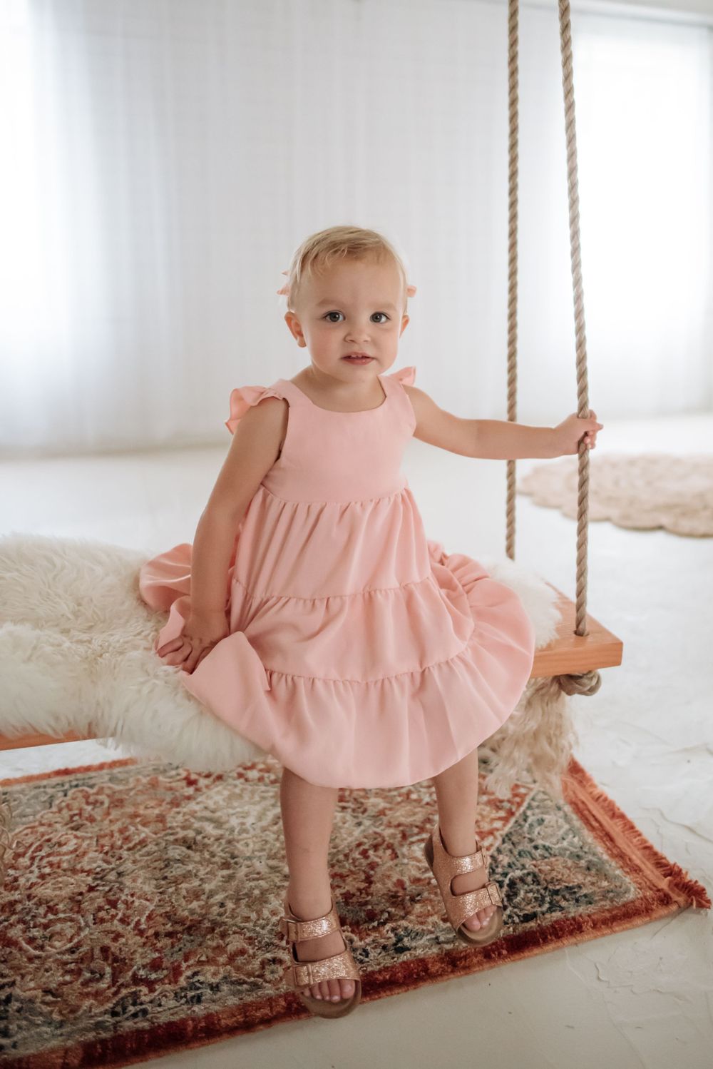 A toddler in pink ruffle dress stands on indoor rope swing against white backdrop.