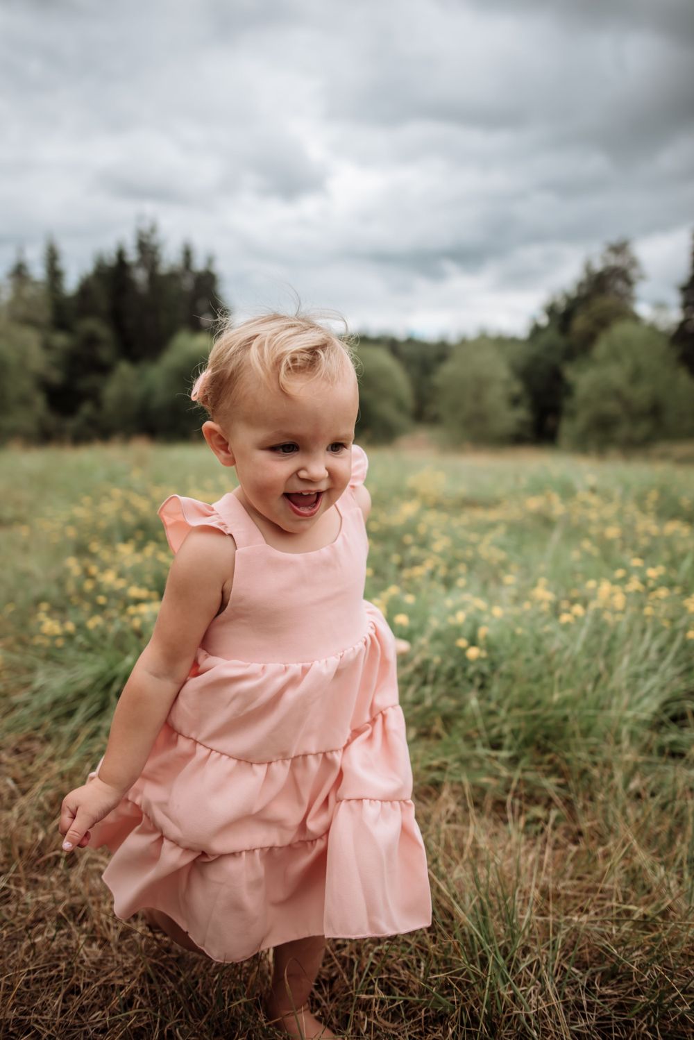 A small child in pink dress plays in field of wildflowers under cloudy sky.