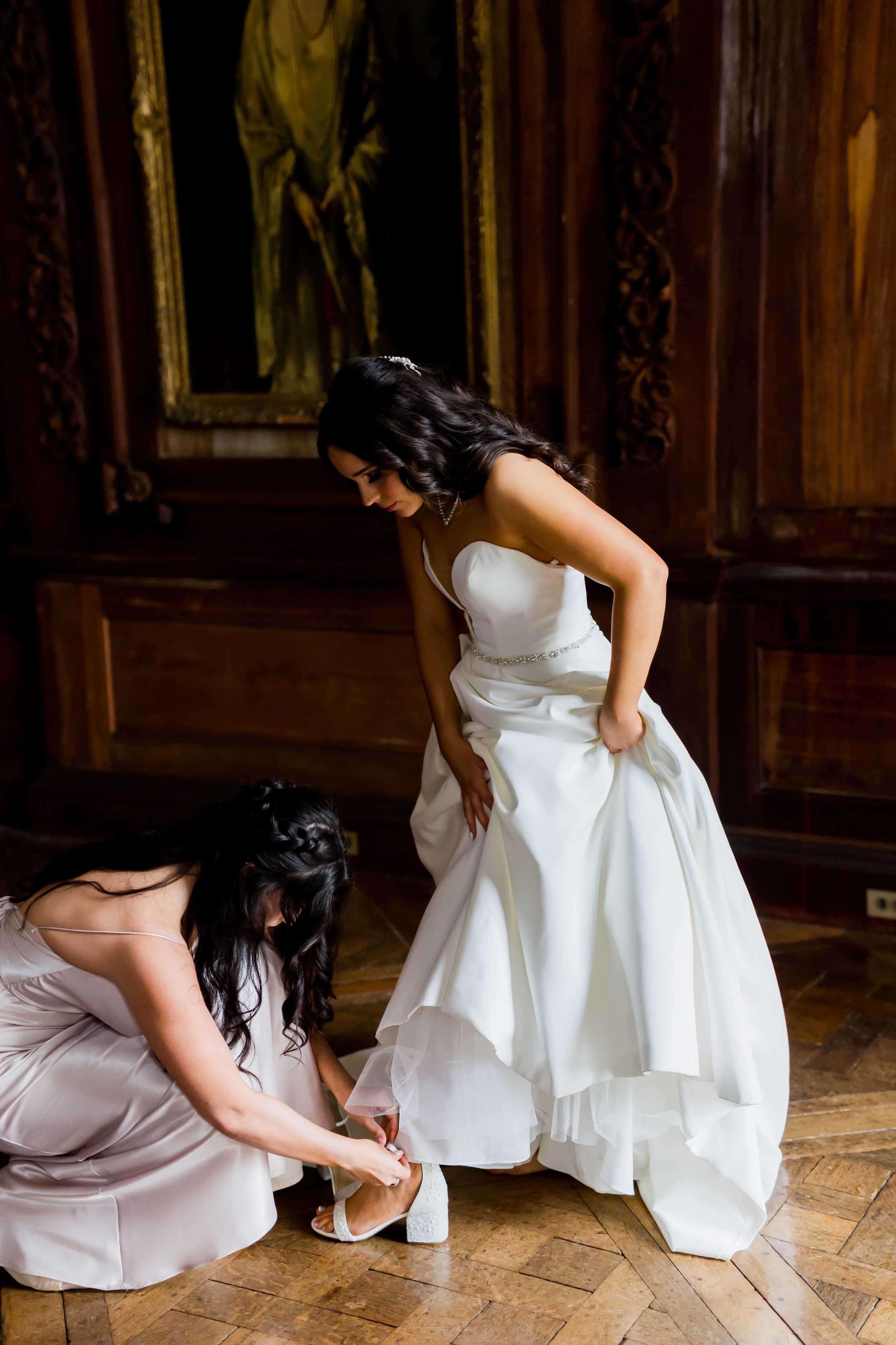 An elegant bride in a white strapless gown gets assistance with her shoes on a parquet floor in a grand room.